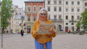 Senior stylish tourist granny woman walking along street, looking for way using paper map in old town. Elderly traveler grandmother getting lost in big city trying to find route. Summer vacation trip