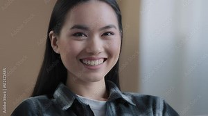 Close-up pensive calm female face Asian 30s woman looking away turning head to camera smiling toothy with perfect white teeth brunette Korean girl with beautiful skin natural make-up indoors posing