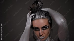 A beautiful young woman with silver paint on her skin, face and hair poses in a studio with a silver metal chain.