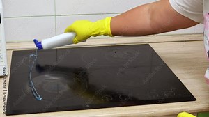 Hands of woman in yellow rubber gloves for cleaning open cap of bottle of cleaning gel for ceramic electric stove. Process of applying cleaning agent to stove