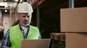 Project Leader is Standing with Laptop Computer and Checking Work Data. Engineers Work Around Him. Factory Worker Engaged in Project Development Using a Laptop Computer