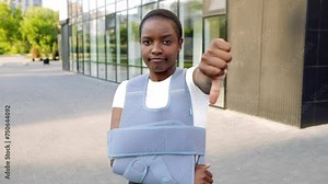 Young african american woman standing outside, broken arm clavicle fracture showing dislike thumb down gesture. Smiling beautiful girl patient near clinic hospital wear bandage arm brace relive pain