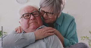 Happy senior couple at home. Old woman hugs man sitting on sofa. Mature grey haired lady hugs white haired husband from behind, talks to him, smiles, tells to have rest and relax. Love, care concept