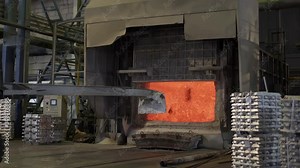 Loader mixes molten aluminum in a blast furnace. Aluminium foundry furnace loaded with metal. Red hot flames glowing and liquid melting. Fire melts aluminum ingots in a blast furnace.