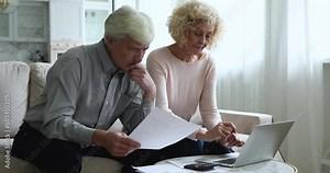 Focused retired senior wife and husband sit on sofa at home paying monthly utility bills, taxes online use laptop, make internet payment via e-bank. Family finances management, budget control concept