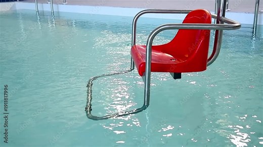 A red plastic wheelchair with a lifting mechanism for the disabled on the background of an indoor pool with blue water.
