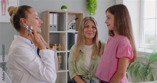 Pediatrician using stethoscope to listen to heartbeat, check health of little girls lungs on clinic examination. Happy child standing in front of doctor and mother for diagnostics, disease prevention