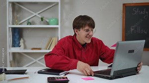 A young guy with glasses communicates using a laptop and smiles while sitting at a table indoors at home. The guy pulls an open laptop towards him and takes off his glasses