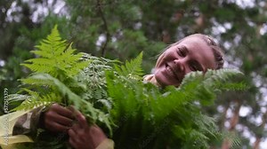 A young woman in a yellow raincoat walks through the forest, breathes clean air. Portrait of a woman in the forest during the rain. In the hands of a woman are green leaves of a fern.