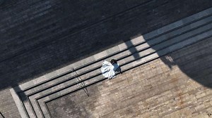 Bride in white dress lies on urban stone stairs casting shadow, groom in black suit holds her tightly on modern square