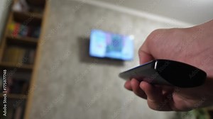 Close-up of a male hand with a TV remote control switches TV channels and turns off the TV. A man is resting on the bed and watching TV.