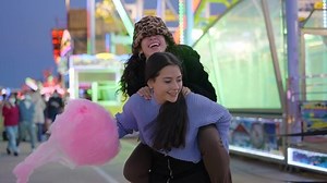 One girl gives another a piggyback ride holding pink candy floss. Best friends teenagers having fun at the funfair at night - slow-motion