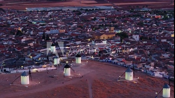 Closer aerial view of illuminated windmills at dusk in Campo de Criptana, Spain, with traditional village rooftops, historic church, and dry landscape of La Mancha in the background