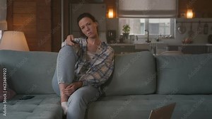 alone frustrated young woman sits on sofa at home and holds tv remote, changing channels monotonously. looks bored, uninterested and depressed.