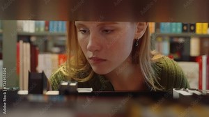 Close-up young woman with red hair and freckles intently browsing through files or books, surrounded by bookshelves in library or coworking space. Education and learning concept