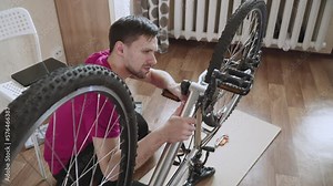 Caucasian thin man uses force to remove crank arm with pedal from aluminum frame mountain bike bottom bracket during home repair. Second connecting rod falls, having lost its support.