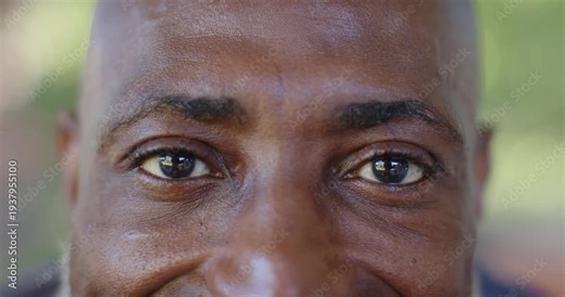 Mature African man holding eye contact, camera reflection causing blinking and smiling in park