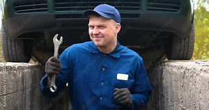 A cheerful car repairman on the street repairs the engine and lower part of the car, shows different emotions, dances with a big wrench. Concept of: Service, Joy, Slow motion, Mood, Happy, Repair.
