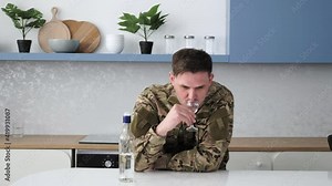 Young caucasian man civil soldier sitting at home kitchen bar counter with bottle and glass drinking vodka alcohol beverage feeling depressed upset desperate lonely after returning home from war.
