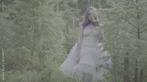 Beautiful woman in white dress and purple hair falling down on the ground in forest - fairytale scene. Side view video of sensual beauty on green grass in slow motion.