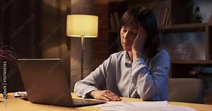 Weary woman continues work at home. Her eyes show signs of fatigue, yet determination remains steadfast. The room is illuminated by the soft glow of lamp.