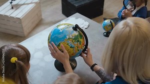 Top view. A stylish teacher with a tattoo shows the kids a globe. Teacher discussing over a globe with kids in geography classroom at elementary school