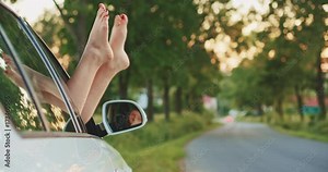 Woman showing legs from the car window and moving them playfully. Slow motion. Filmed in 4K DCi resolution. Young woman relaxing in the car during a stop on a road trip, with sunlit road background.