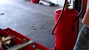 Mechanics garage slow pan up from red tool box with air compressor tools and oil changing equipment in focus, as mechanic is out of focus in background working on car on lift Premium Stock Video Footage