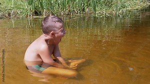 A Sleepy Yawning Child Sits in a Lotus Position in a Shallow River at Sunset. Tired, resting teenager relaxes in the water in the rays of sunlight, against the background of green reeds in the lake.