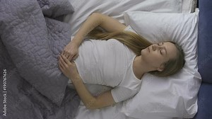 Top view of young pretty woman lying on her back in bed and snoring while sleeping. Tired female in white t-shirt fell asleep late in bed and snores with open mouth.