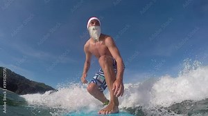 SLOW MOTION CLOSE UP: Cheerful Santa surfer riding fun waves in sunny ocean. Smiling surfer Santa Clause surfing waves towards the shore to wish a Merry Christmas. Funny santa Clause surfing