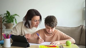 Online remote: mom helping son to do homework and smiling while sitting at table in home spbd. Closeup view of smart boy writes in notebook and studies, his mother helps him, looks with smile and sits
