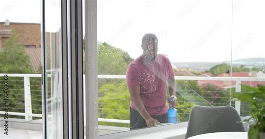 Adult male noticing streaks, spraying bottle and squeegeeing glass door on balcony, removing marks