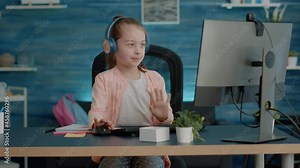 Little child waving at video call camera while wearing headphones for online class lesson with teacher. Young girl talking on video conference on computer for remote education courses