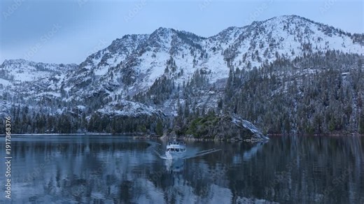 Aerial drone view of tourist cruise boat moving across calm Lake Tahoe water near rocky island and snow covered pine forest shoreline. Winter alpine landscape. Lake Tahoe, California, USA.