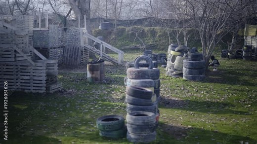 An outdoor paintball field features wooden obstacles and tire barricades. A player is visible, actively participating in the game.
