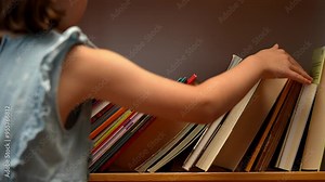 A young schoolgirl walks up to a bookshelf filled with books and pulls out one book from between the others.