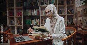 Female muslim student is studying at desk in library, using laptop and books. Girl wearing hijab is preparing for exams - student lifestyle, modern islam concept 4k close up