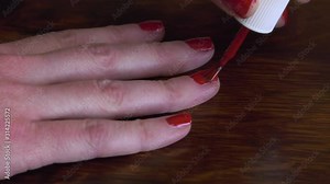 Four fingers of healthy right hand close-up, woman painting fingernails with eye-catching bright red nail polish second coat, on oak wood desktop.