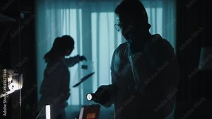 Forensic specialists work at the crime scene, in a dark apartment lit by blue red light from police sirens. A man with a flashlight speaks on his smartphone, while a woman examines the murder weapon. Stock Video
