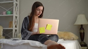 A beautiful woman works remotely using a laptop while sitting on the bed in her cozy bedroom. Sticky notes on the lid of a laptop as a method of systematic planning