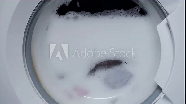 Washing clothing in domestic washing machine. Close up video of spinning washing machine drum with foam spraying door