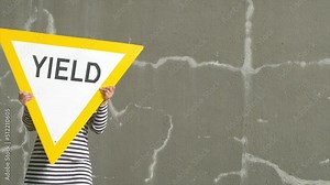 Person carries yield road sign from right to left. Bright yellow against cracked gray wall. Eye-catching symbol for abstract concepts: give way, change, concede, profit, revenue, crop, and harvest. Stock Video