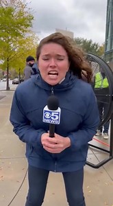 Reporter Claims ‘100 MPH Winds’—Camera Accidentally Reveals the Real Storm Behind the Storm” At first glance, the live shot looks dramatic—an on-scene reporter gripping her microphone with both hands, hair whipping sideways, voice straining over the “violent gusts” she insists are “making it nearly impossible to stand.” Her urgency feels genuine… until viewers notice two tiny details the camera operator apparently didn’t. Just over her shoulder, a couple strolls by at a perfectly casual pace, pu