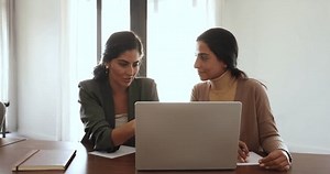 Business application developer woman presenting software on laptop to female customer, showing app work. Two businesswomen talking at computer in office, speaking, listening, nodding