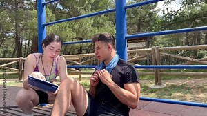 video of portrait of a personal trainer woman explaining the exercise table to train a young man while he is resting sitting on the floor with the towel around his neck