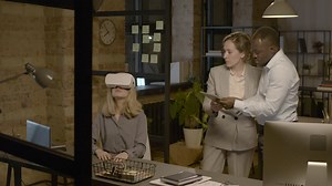 Free stock video - Woman at desk in the office wearing virtual reality glasses while a woman and man is controlling her with tablet