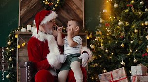Little boy sitting on Santa Claus lap listening to his story, next to the Christmas tree in room with festive interior. Shooting in slow motion. Stock Video