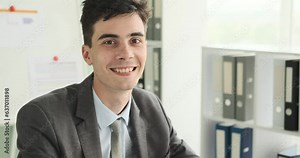 Smiling businessman is seen looking directly into the camera. With a friendly and approachable smile, he exudes confidence and positivity. The office setting reflects a professional environment.