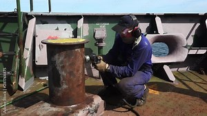 RIGA/LATVIA - JUNE 24 2019: specially equipped worker removes rust from vessel anchor stand with angle grinder on sailing ship deck on June 24 in Riga